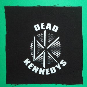 May include: Black fabric patch with white text and a white graphic. The text reads "DEAD KENNEDYS" and the graphic is a stylized starburst.
