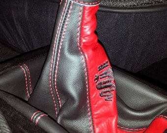 Puede incluir: Una funda de palanca de cambios de cuero negro con costuras rojas y un inserto de cuero rojo. El inserto tiene la palabra "Alfa" bordada.