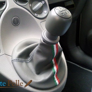 Puede incluir: Funda de palanca de cambios de cuero gris con una perilla de palanca de cambios gris y una franja verde, blanca y roja. La perilla de palanca de cambios tiene los números 1, 2, 3, 4, 5 y R.