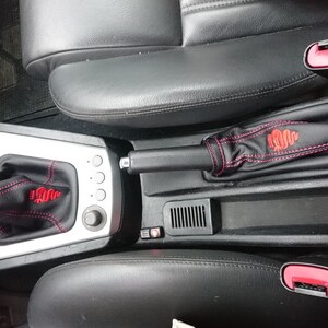 Puede incluir: Primer plano del interior de un coche con una funda de palanca de cambios de cuero negro y una funda de freno de mano de cuero negro. Ambas fundas tienen costuras rojas y un logotipo de Alfa Romeo bordado en rojo.