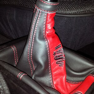 Può includere: Un soffietto del cambio in pelle nera con cuciture rosse e un inserto in pelle rossa. L'inserto ha la parola "Alfa" ricamata.