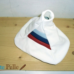 Può includere: Fodera per leva del cambio in pelle bianca con strisce rosse, blu e blu chiaro. La fodera ha la forma di un cono con una base larga e una cima stretta. La fodera è appoggiata su una superficie di legno. Il testo "L'Arte Pelle" è visibile nell'angolo in basso a sinistra dell'immagine.