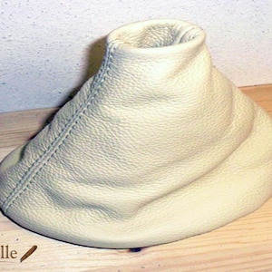 May include: A cream-colored leather gear shift boot with a stitched seam. The boot is sitting on a wooden surface.