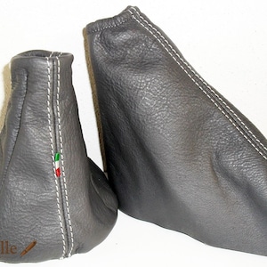 Puede incluir: Dos fundas de cuero grises para palanca de cambios de coche con costuras blancas. Una funda tiene un diseño alto y cónico, y la otra es rectangular. La bandera italiana está cosida en el lateral de la funda cónica.