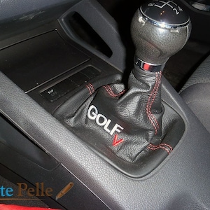 Könnte beinhalten: Schwarzer Lederschalthebelbalg mit roten Nähten und "GOLF V" Stickerei an der Seite.