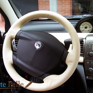 Può includere: Primo piano di un volante in pelle beige all'interno di un'auto. Il volante ha un centro nero con un logo argentato e la scritta "Airbag" stampata su di esso. Il volante è circondato da un cruscotto nero con vari controlli e indicatori.