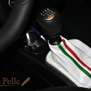 Pode incluir: Botão de cambio de couro branco com listras verdes e vermelhas. O botão está instalado na alavanca de câmbio de um carro.