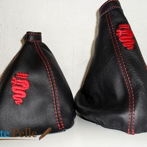 Op de afbeelding: Twee zwarte leren versnellingspookhoezen met rode stiksels en een rood geborduurd Alfa Romeo logo.
