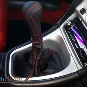 Może przedstawiać: Zbliżenie na skórzaną gałkę zmiany biegów w samochodzie w kolorze czarnym z czerwonymi szwami. Gałka jest zamontowana w konsoli środkowej samochodu.