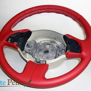 Puede incluir: Volante de cuero rojo con costuras blancas. El volante está retirado de un coche y el cubo central es visible.
