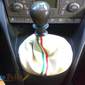 Puede incluir: Funda de palanca de cambios de cuero blanco con rayas verdes, blancas y rojas. La funda está instalada en un coche con interior negro.