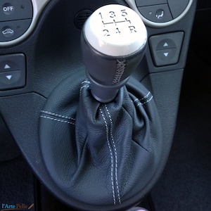 Puede incluir: Primer plano de una funda de palanca de cambios de cuero negro con costuras blancas. La perilla de la palanca de cambios es blanca con números negros 1, 2, 3, 4, 5, R y una funda de cuero negro.