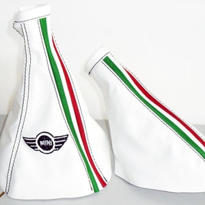 Puede incluir: Dos fundas de palanca de cambios de cuero blanco con rayas verdes y rojas. La funda de la izquierda tiene un logotipo Mini bordado en negro con alas.