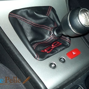 May include: Close-up of a black leather gear shift boot with red stitching and a silver gear shift knob with a 6-speed pattern. The boot has the word "S.S." embroidered on it. The image also shows a red hazard light button and other buttons on the center console.