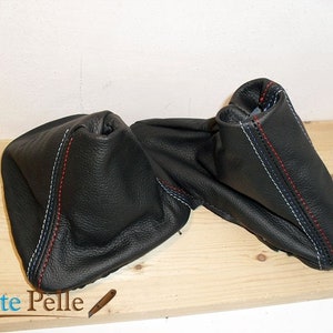 Könnte beinhalten: Schwarze Lederschaltmanschetten mit roten und weißen Nähten. Die Bezüge bestehen aus einem strukturierten Ledermaterial und sind für den Schalthebel eines Fahrzeugs konzipiert. Das Bild zeigt zwei Bezüge, einer davon ist teilweise geöffnet.