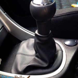 May include: Close-up of a black leather gear shift boot with a silver gear knob. The boot is stitched with white thread and has a smooth, textured surface.