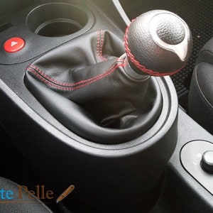 Puede incluir: Primer plano de una palanca de cambios de coche de cuero negro con costuras rojas. La palanca tiene un acento plateado y está rodeada por una funda de cuero negro.