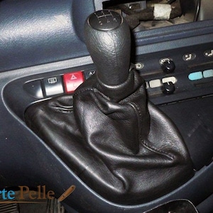May include: Close-up of a black leather gear shift boot and knob in a vehicle's interior. The shift knob has a 5-speed pattern. The image also shows the dashboard with various controls and the text "l'Arte Pelle".