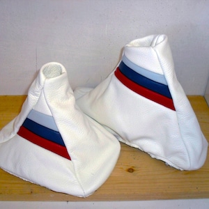 Può includere: Due soffietti per leva del cambio in pelle bianca con strisce rosse, blu e grigie. I soffietti sono su una superficie di legno.