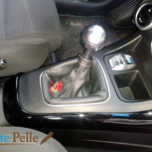 Könnte beinhalten: Nahaufnahme eines schwarzen Lederschalthebels mit roten Nähten. Der Schalthebel ist in der Mittelkonsole eines Autos installiert, die ebenfalls schwarz ist. Der Schaltknauf ist silberfarben und hat eine schwarze Basis.