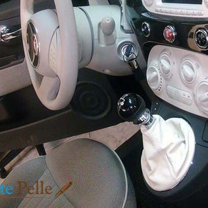 May include: White leather gear shift boot with a black knob in a car interior. The car has a black dashboard with white buttons and a black steering wheel.