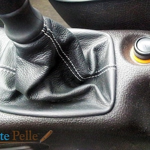 May include: Close-up of a black leather car gear shift boot with white stitching. The boot is installed in a car's center console. A black button with an orange ring is visible to the right of the boot.