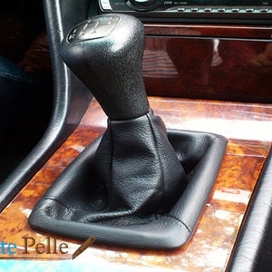 Könnte beinhalten: Nahaufnahme eines schwarzen Lederschalthebels mit einem schwarzen Schaltknauf. Der Schaltknauf hat eine silberne Einfassung und einen schwarzen Lederschuh. Der Schalthebel befindet sich in einem Auto mit einer Holzverkleidung.