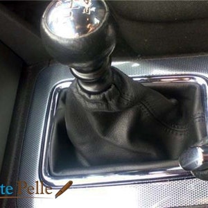May include: Close-up of a black leather gear shift boot with a chrome trim around the base. The gear shift knob is silver and has a black leather cover.