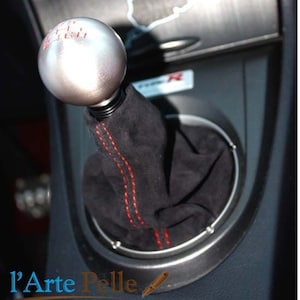 Peut inclure: Un soufflet de levier de vitesses en cuir noir avec des coutures rouges et un pommeau de levier de vitesses argenté avec des chiffres rouges. Le pommeau est rond et a une surface lisse.