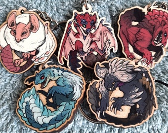May include: Five wooden keychains featuring different monster designs from the Monster Hunter video game series. The keychains are painted in vibrant colors, including red, blue, white, and black. Each keychain features a different monster, including a red dragon, a blue lizard, a white furry creature, a black and white monster with spikes, and a red monster with sharp teeth.