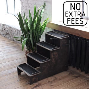 Puede incluir: Un escalón de madera marrón oscuro para mascotas con tres escalones, cada uno con una superficie negra texturizada. Los escalones están diseñados con una estética simple y funcional. La imagen también incluye una planta en maceta y el texto "NO EXTRA FEES".