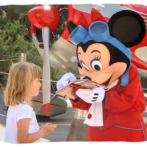 K&ouml;nnte beinhalten: Ein rotes und wei&szlig;es Flugzeug mit einer Mickey-Maus-Figur, die eine rote Jacke und einen blauen Schal tr&auml;gt. Die Figur signiert ein Autogramm f&uuml;r ein junges M&auml;dchen mit blonden Haaren.