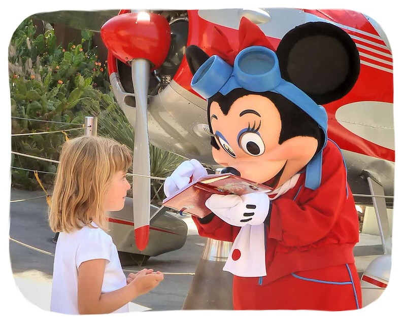 K&ouml;nnte beinhalten: Ein rotes und wei&szlig;es Flugzeug mit einer Mickey-Maus-Figur, die eine rote Jacke und einen blauen Schal tr&auml;gt. Die Figur signiert ein Autogramm f&uuml;r ein junges M&auml;dchen mit blonden Haaren.