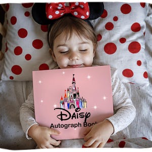 Könnte beinhalten: Ein Kind mit Minnie Mouse-Ohren hält ein rosa Disney-Autogrammbuch. Das Buch zeigt eine farbenfrohe Schlossillustration und das Wort "DAISY" über "Autograph Book". Das Kind liegt in einem Holzbett mit einem Kissen mit Tupfen.