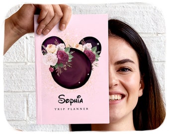 May include: A pink planner with a black and purple Mickey Mouse head design with floral accents. The planner says "Sophia Trip Planner" on the front.