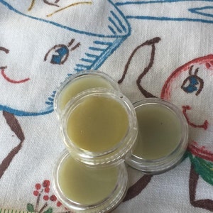 May include: Three small clear containers filled with a light green balm. The containers are on a white fabric with a red and green cartoon design. The fabric has the text "Rock 'n' Roll" embroidered on it.