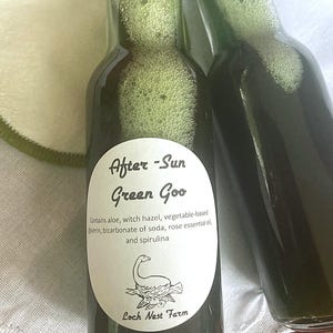 May include: Two glass bottles filled with a green liquid, labeled "After-Sun Green Goo". The label also lists ingredients including aloe, witch hazel, vegetable-based glycerin, bicarbonate of soda, rose essential oil, and spirulina. The label has a drawing of a Loch Ness Monster.