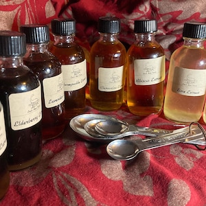 May include: A collection of glass bottles with black caps, each labeled with a different extract flavor, such as Elderberry, Lavender, and Lime. Silver measuring spoons are arranged in front of the bottles on a red patterned surface.