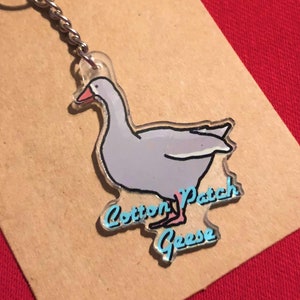 May include: A clear acrylic keychain featuring a cartoon goose with a pink beak and blue eyes. The goose is walking and the text "Cotton Patch Goose" is written in blue on the keychain.