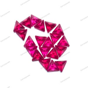 May include: A collection of triangular, faceted, hot pink gemstones. The gems are arranged in a loose cluster against a white background. The gems have a vibrant, saturated color and a glossy finish. The brand name "Gemsnjewele" is printed around the image.