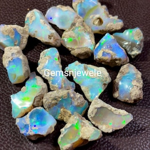 Op de afbeelding: Een verzameling ruwe, natuurlijke opaal edelstenen. De opalen zijn een mix van wit, grijs en bruin met flitsen van groen, blauw en iriserende kleuren. De opalen zijn onregelmatig van vorm en grootte.