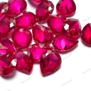 Pode incluir: Uma coleção de pedras preciosas de rubi facetadas, em forma de coração e de pêra. As gemas são de uma cor vermelho-rosa vibrante e estão dispostas sobre um fundo branco. As facetas capturam a luz, criando um efeito brilhante.