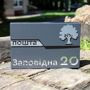 Può includere: Una cassetta postale grigio scuro con la parola "ПОШТА" e l'indirizzo "Заповідна 20" ritagliati. Un disegno ad albero argentato è sul lato destro. La cassetta postale è rettangolare e poggia su una superficie di legno.