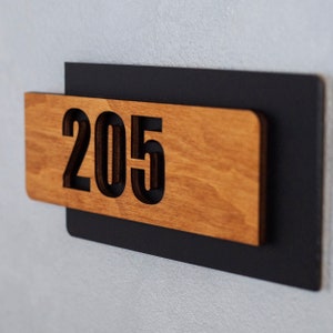 Puede incluir: Un letrero de madera con el n&uacute;mero 205 recortado en letras negras. El letrero est&aacute; montado sobre un fondo negro.
