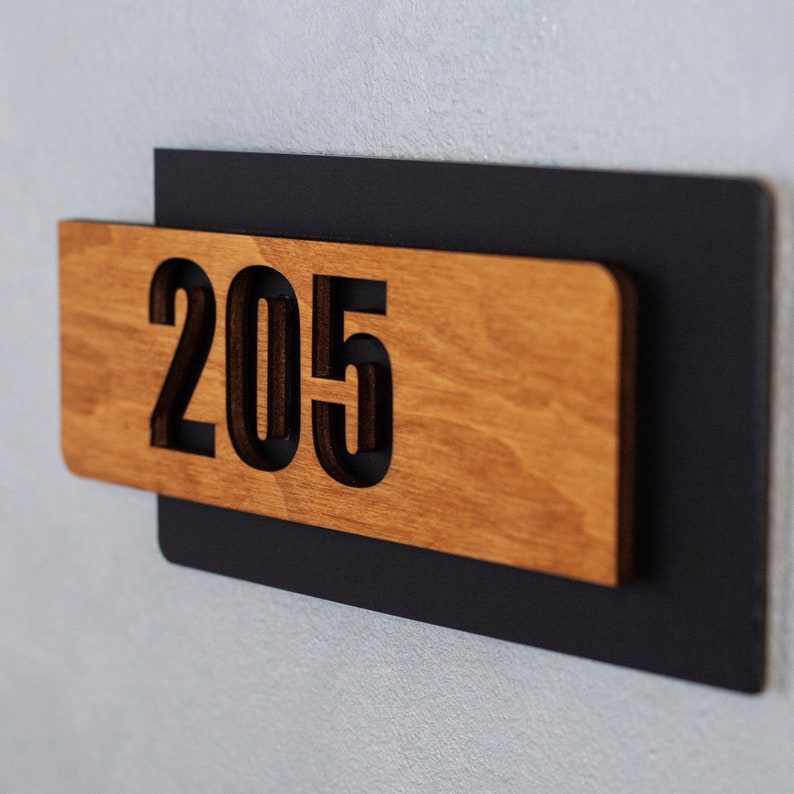 Puede incluir: Un letrero de madera con el n&uacute;mero 205 recortado en letras negras. El letrero est&aacute; montado sobre un fondo negro.