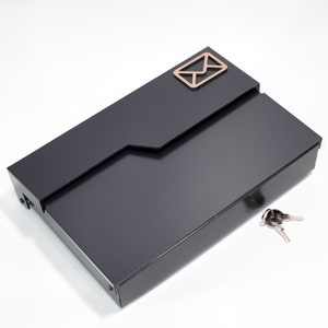 Puede incluir: Un buzón de metal negro con una ranura para el correo y una cerradura. El buzón tiene un pequeño icono de sobre en la parte delantera.