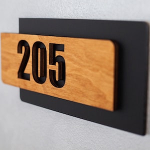 Puede incluir: Un letrero de n&uacute;mero de casa de madera con el n&uacute;mero 205 recortado. El letrero est&aacute; montado sobre un fondo negro.