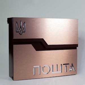 Puede incluir: Un buzón de color bronce con un acabado de metal cepillado. El buzón presenta un acento marrón oscuro y un diseño recortado con la palabra "ПОШТА". El escudo de armas ucraniano se muestra en la esquina superior izquierda.