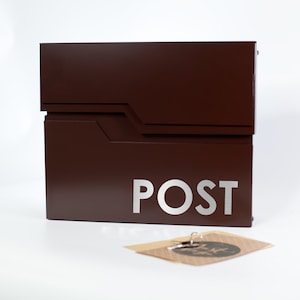 Op de afbeelding: Een moderne, bruine brievenbus met het woord "POST" in zilveren letters. De brievenbus heeft een geometrisch ontwerp en een gleuf voor post. Een kleine kaart met sleutels ligt voor de brievenbus.