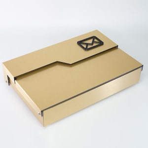 May include: A gold-colored, rectangular box with a black envelope icon on the top flap. The box has a brushed metal finish and black trim, suggesting a sleek and modern design. The flap is slightly open, revealing the interior.